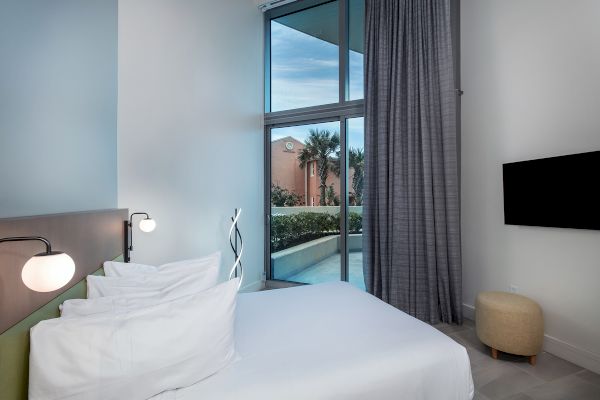 A modern bedroom with a bed, pillows, lamps, a TV on the wall, and a window with curtains overlooking an outdoor area with plants.