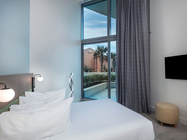 A modern bedroom with a bed, pillows, lamps, a TV on the wall, and a window with curtains overlooking an outdoor area with plants.