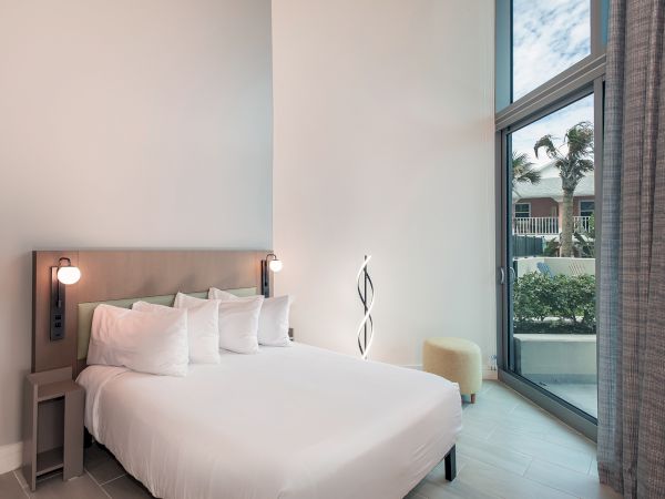 A modern bedroom with a double bed, bedside tables, and lamps. A large window offers an outdoor view with greenery, and there's a unique floor lamp.