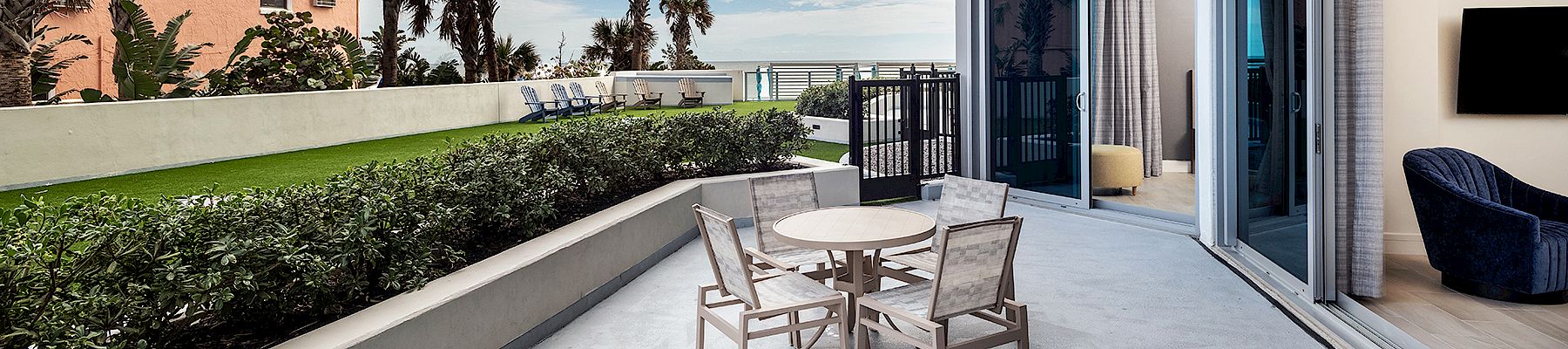 A spacious balcony with a table and chairs, overlooking a garden and ocean view, adjacent to a modern room with large windows and sliding doors.