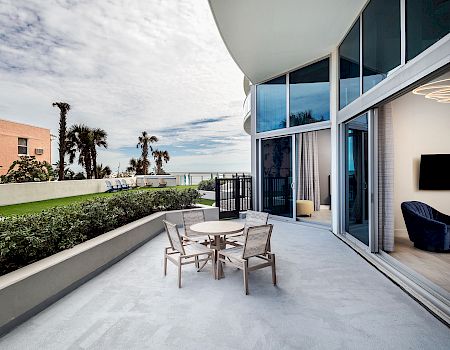 A spacious balcony with a table and chairs, overlooking a garden and ocean view, adjacent to a modern room with large windows and sliding doors.