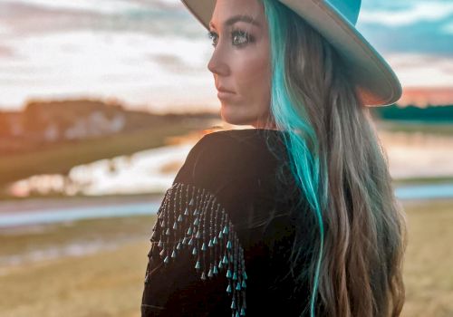 A person stands outdoors wearing a hat and a beaded jacket with long hair, gazing into the distance near a scenic water body at sunset.