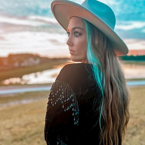 A person stands outdoors wearing a hat and a beaded jacket with long hair, gazing into the distance near a scenic water body at sunset.