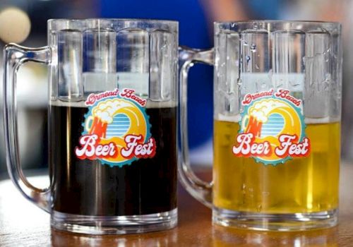 Two clear beer mugs labeled &ldquo;Grand Best Beer Fest,&rdquo; one filled with dark beer and the other with golden beer, sitting on a table.
