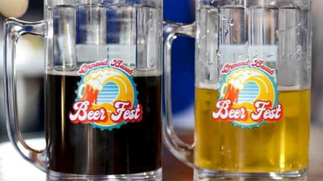 Two clear beer mugs labeled &ldquo;Grand Best Beer Fest,&rdquo; one filled with dark beer and the other with golden beer, sitting on a table.