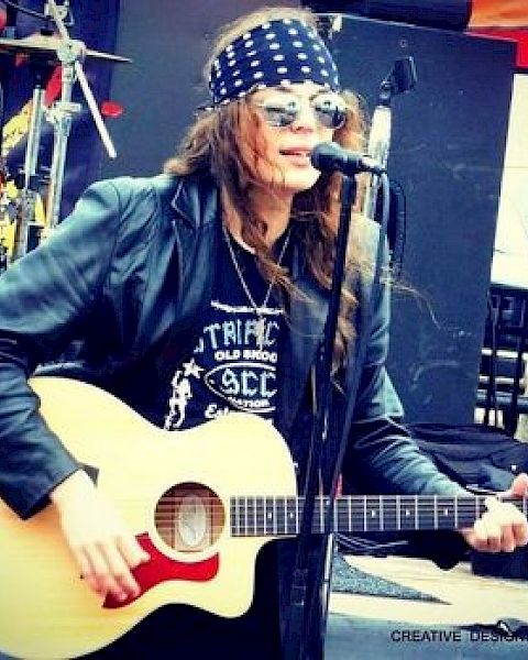 A person wearing a bandana and sunglasses is playing an acoustic guitar and singing into a microphone on an outdoor stage.