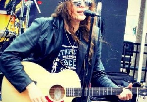 A person wearing a bandana and sunglasses is playing an acoustic guitar and singing into a microphone on an outdoor stage.