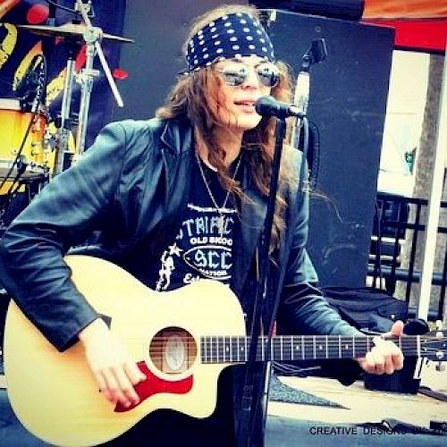 A person wearing a bandana and sunglasses is playing an acoustic guitar and singing into a microphone on an outdoor stage.