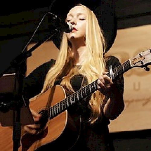 A person with long blonde hair is playing an acoustic guitar and singing into a microphone on stage, framed by a dark background.