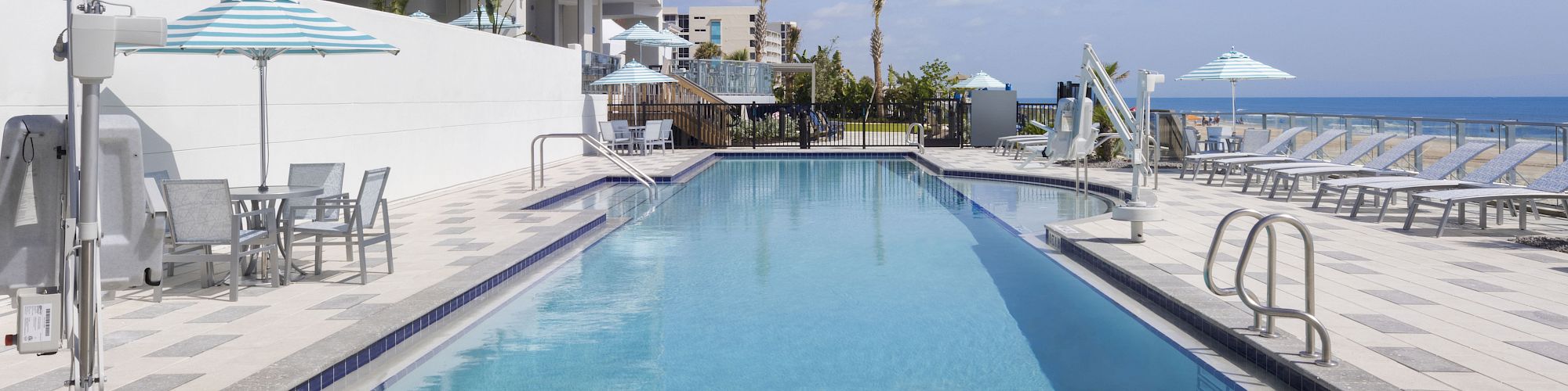 The image shows a modern building with a clear sky, a long pool, and a hot tub in the foreground.