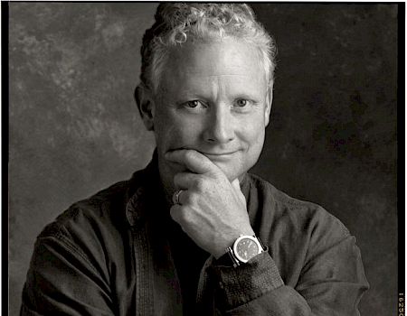 A black-and-white photo of an unidentified person with curly hair, wearing a watch, and posing with one hand under their chin.