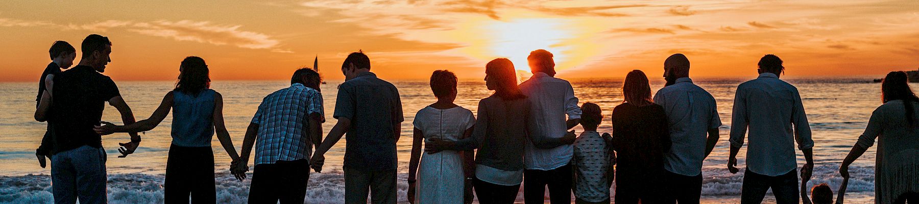 A group of people standing on a beach, holding hands, facing a sunset over the ocean with a cloudy sky.