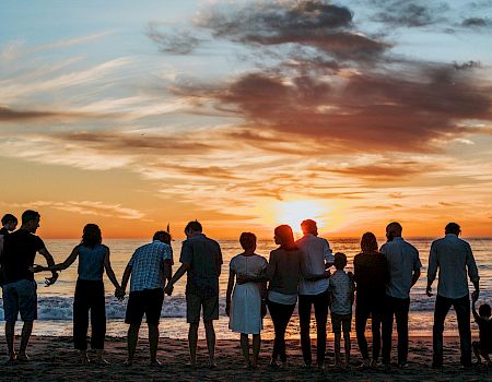 A group of people standing on a beach, holding hands, facing a sunset over the ocean with a cloudy sky.