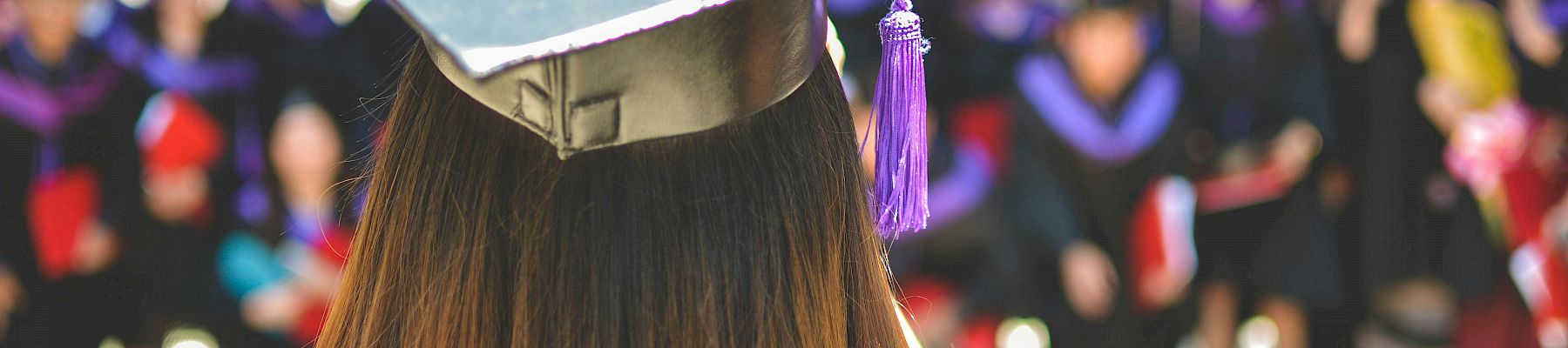 A person in a graduation cap and gown stands facing a crowd of similarly dressed individuals, suggesting a graduation ceremony.