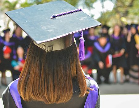 A person in a graduation cap and gown stands facing a crowd of similarly dressed individuals, suggesting a graduation ceremony.