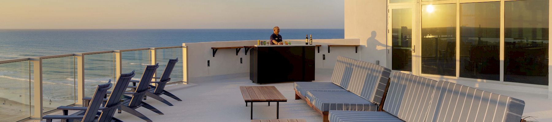 A rooftop terrace with ocean views, featuring lounge chairs, striped cushioned seating, tables, and a bar area with a person standing behind it.