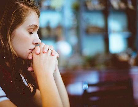 A woman with closed eyes is clasping her hands together while sitting in what appears to be a peaceful setting.