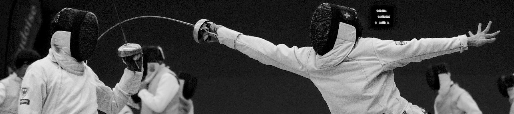 Two fencers in action during a match, wearing protective gear and masks, swords crossed, with others visible behind them.