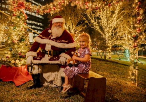 A child in a purple dress sits beside Santa Claus in a festive outdoor setting with decorative lights and a wreath.
