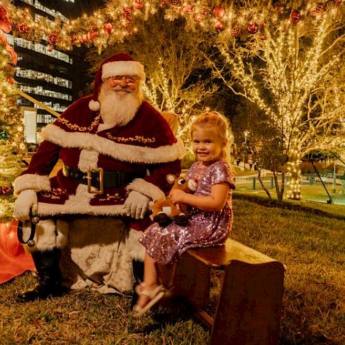 A child in a purple dress sits beside Santa Claus in a festive outdoor setting with decorative lights and a wreath.