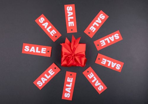 A red gift box with a bow is surrounded by red “SALE” tags arranged in a circular pattern on a black background.