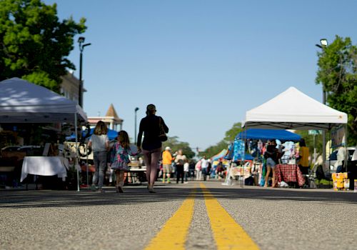 A street market scene with people walking, stalls set up on both sides, a clear sky, and trees and buildings in the background.