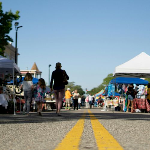 A street market scene with people walking, stalls set up on both sides, a clear sky, and trees and buildings in the background.