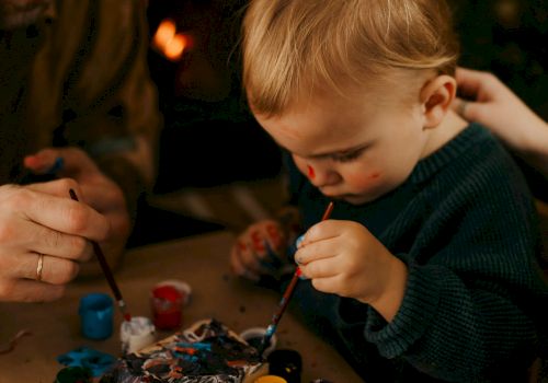 An adult and a young child are painting together at a table, with various paint containers and brushes in front of them, near a cozy fireplace.