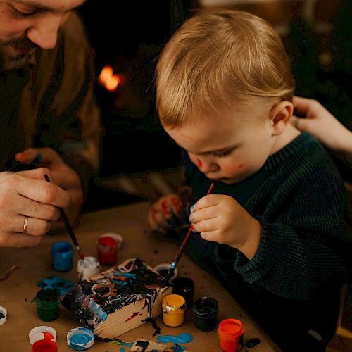 An adult and a young child are painting together at a table, with various paint containers and brushes in front of them, near a cozy fireplace.