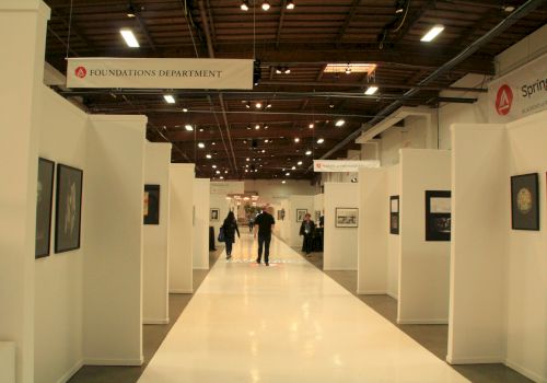 A spacious art gallery with white walls displays framed artworks, and two people walk down the central aisle. Signs indicate 