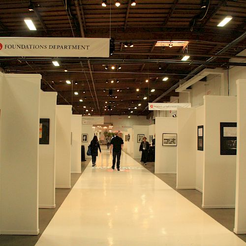 A spacious art gallery with white walls displays framed artworks, and two people walk down the central aisle. Signs indicate 