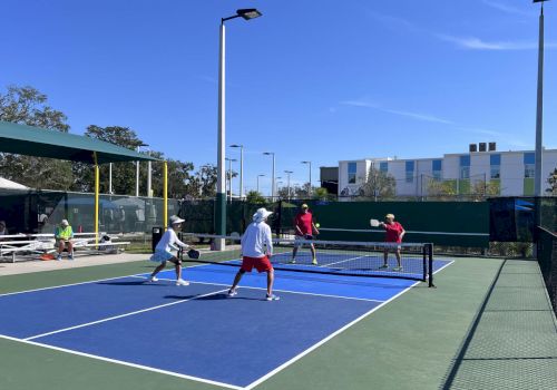 People playing paddle tennis on a bright blue-green court under a clear sky with tall light poles.