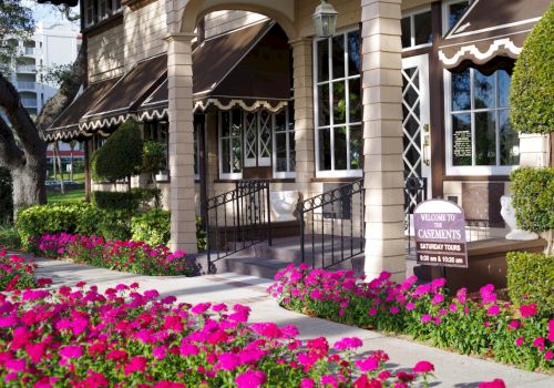 A building with awnings, surrounded by vibrant pink flowers and manicured bushes, features a welcoming sign by the entrance.