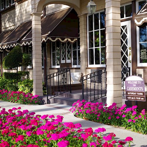 A building with awnings, surrounded by vibrant pink flowers and manicured bushes, features a welcoming sign by the entrance.
