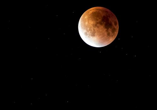 The image shows a lunar eclipse, with the moon appearing reddish-orange against a black sky dotted with stars.