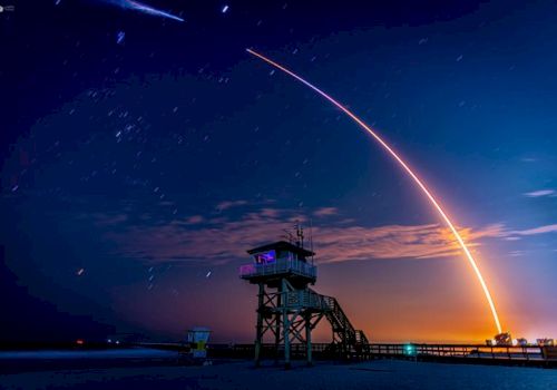 The image shows a long exposure of a rocket launch, creating a bright arc in the sky, with a lifeguard tower in the foreground at dusk.