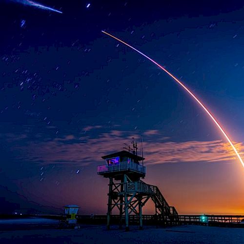 The image shows a long exposure of a rocket launch, creating a bright arc in the sky, with a lifeguard tower in the foreground at dusk.