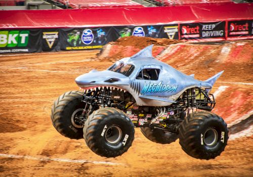 A monster truck designed to look like a shark, performing a jump on a dirt track during a motorsport event with banners in the background.