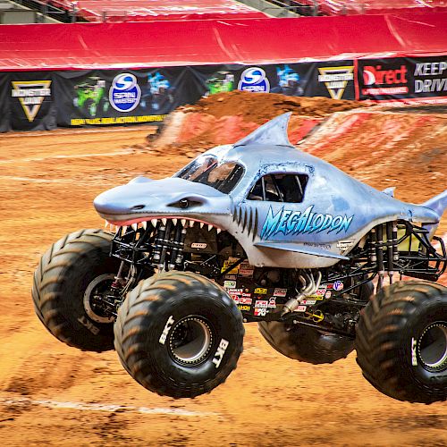 A monster truck designed to look like a shark, performing a jump on a dirt track during a motorsport event with banners in the background.