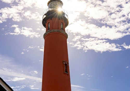 The image shows a tall red lighthouse under a partly cloudy sky, with the sun shining brightly behind its top.