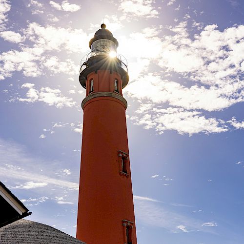 The image shows a tall red lighthouse under a partly cloudy sky, with the sun shining brightly behind its top.