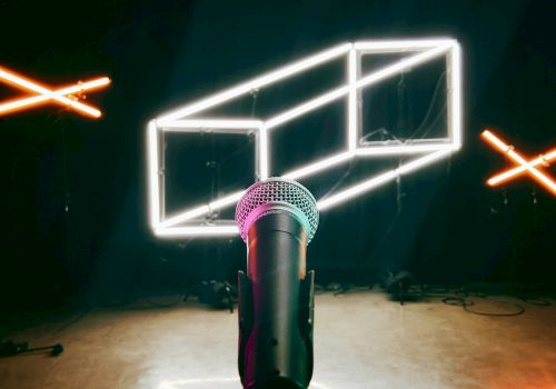 A microphone on a stand is in the foreground, with illuminated geometric neon shapes and X marks in the background, suggesting a stage setup.