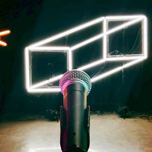 A microphone on a stand is in the foreground, with illuminated geometric neon shapes and X marks in the background, suggesting a stage setup.