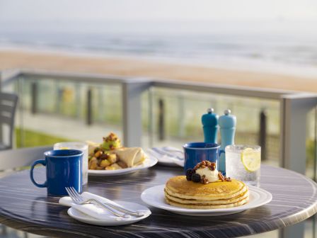 A table setup for breakfast by the sea, featuring pancakes, fruit, blue mugs, cutlery, salt and pepper shakers, and a glass of water with lemon.