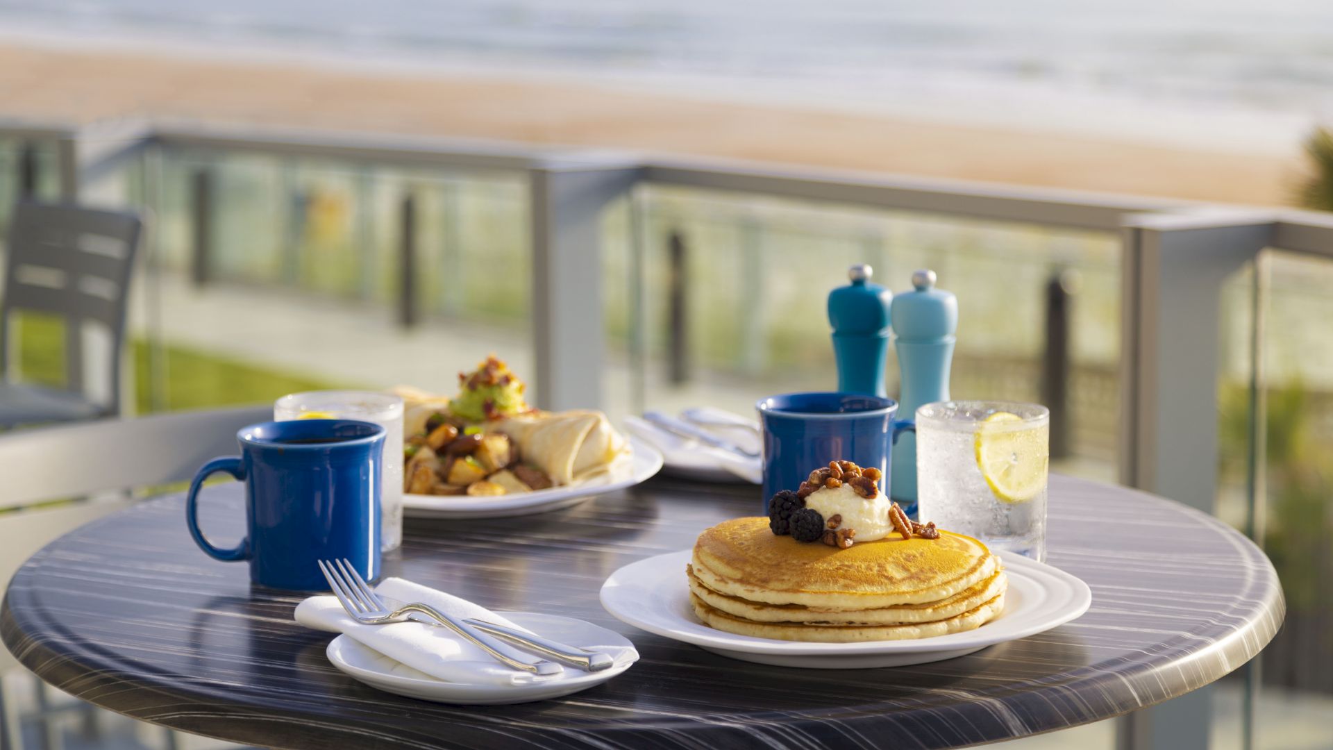 A table set for breakfast with pancakes, coffee, a wrap with potatoes, and a glass of water overlooks a scenic beach view, ending the sentence.