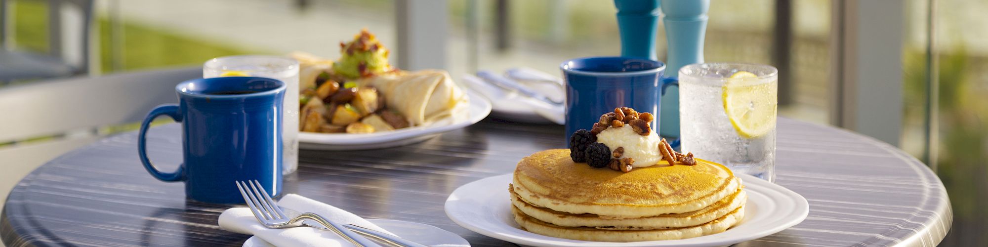 A table set for breakfast with pancakes, coffee, a wrap with potatoes, and a glass of water overlooks a scenic beach view, ending the sentence.