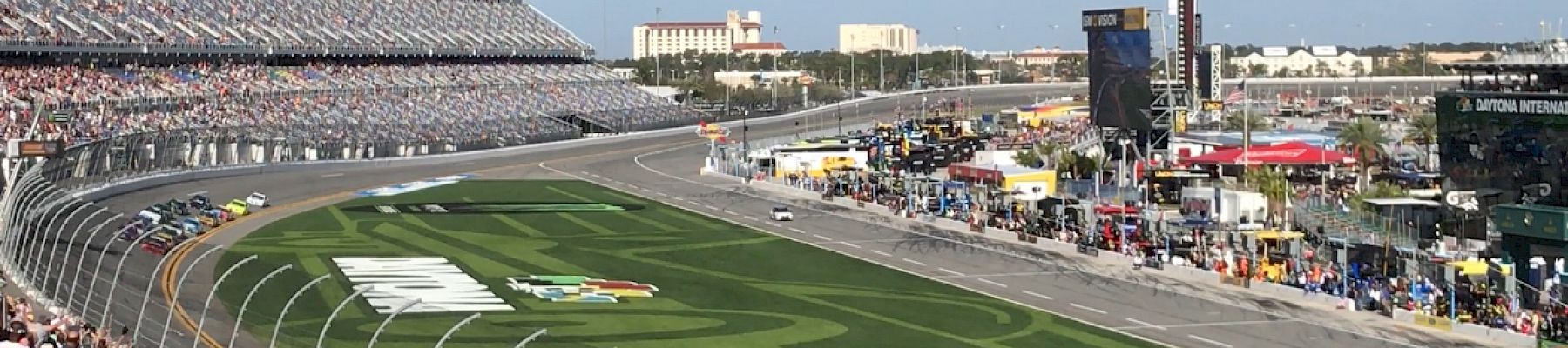 A car racing event is taking place at a track, with cars on the circuit, spectators in the stands, and various facilities and structures in the background.