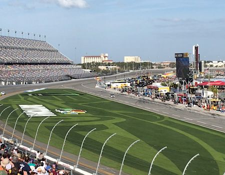 A car racing event is taking place at a track, with cars on the circuit, spectators in the stands, and various facilities and structures in the background.