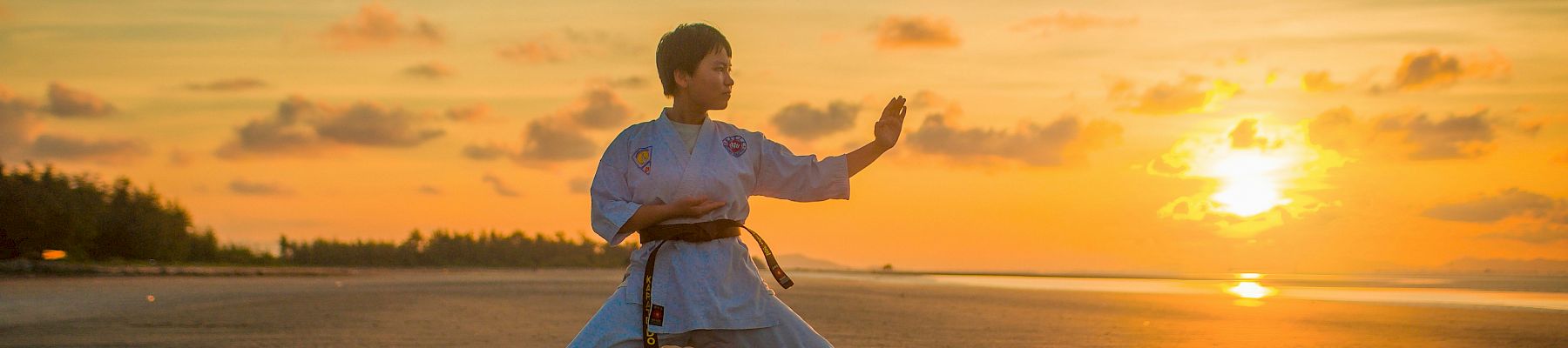 A person dressed in martial arts attire practices a stance on a vast, sandy beach during a beautiful sunset with orange and yellow hues.