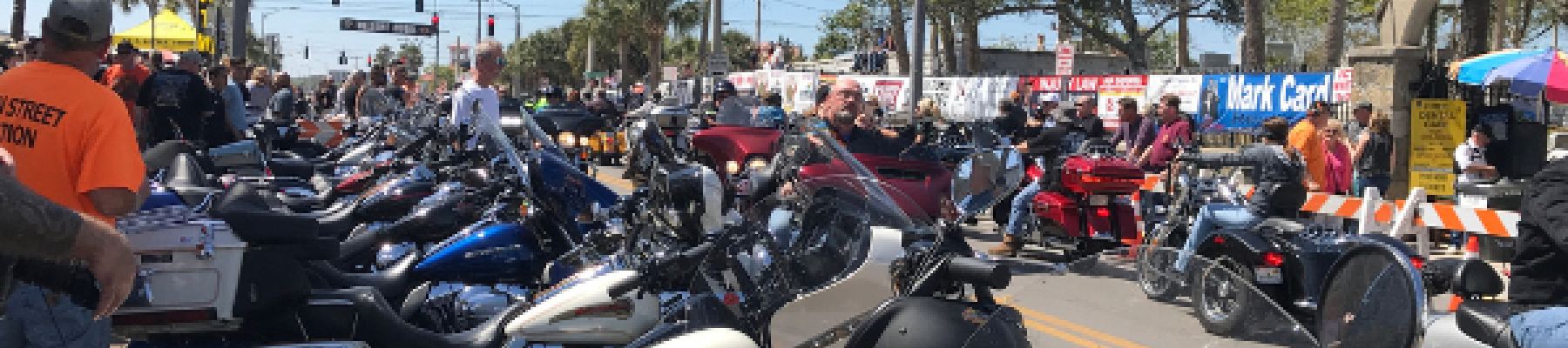 The image shows a large gathering of motorcycles parked along a street, with a banner reading 